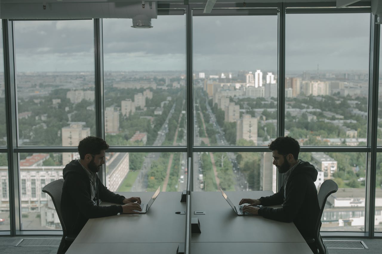 Two men working on laptops in a modern office with a panoramic city view through large windows.
